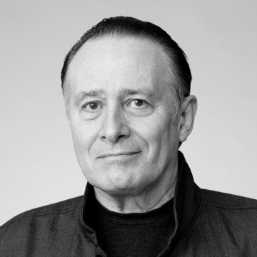 Mustavalkoisessa muotokuvassa poseeraa keski-ikäinen mies, jolla on takapitkät hiukset, tumma takki ja hento hymy, vaaleaa taustaa vasten.
