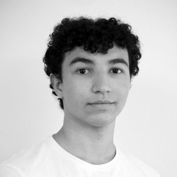 Nuori henkilö, jolla on lyhyet, kiharat tummat hiukset ja neutraali ilme, yllään tavallinen valkoinen paita, poseeraa vaaleaa taustaa vasten.