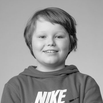 Lyhythiuksinen lapsi hymyilee hellästi, päällään Nike-logolla varustettu huppari. Kuva on mustavalkoinen, ja tausta on tavallinen ja vaalea.