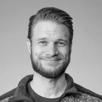 Vaaleahiuksinen, parrakas mies hymyilee kameralle. Hänellä on vetoketjullinen villapaita t-paidan päällä. Tausta on tavallinen ja vaalea. Kuva on mustavalkoinen.