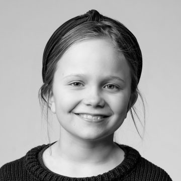 Mustavalkoinen muotokuva nuoresta tytöstä hymyilemässä. Hänellä on yllään tumma otsapanta ja kuvioitu pusero. Hänen hiuksensa ovat hieman sotkuiset, ja tausta on tavallinen ja vaalea.