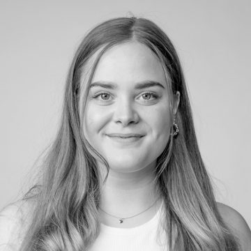 Mustavalkoinen muotokuva nuoresta naisesta, jolla on pitkät, suorat hiukset ja joka hymyilee pehmeästi. Hänellä on yllään vaalea toppi, kaulakoru ja pienet korvakorut, ja hän katsoo suoraan kameraan tavallista taustaa vasten.