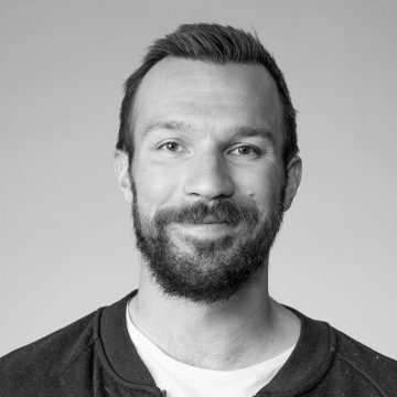 Lyhythiuksinen, parrakas mies, jolla on tumma takki vaalean paidan päällä, hymyilee kameralle vaaleaa taustaa vasten.