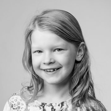 Mustavalkoinen muotokuva nuoresta tytöstä, jolla on pitkät hiukset, hymyilevä ja etuhampaissaan aukko, päällään kuviollinen toppi, taustana tavallinen tausta.