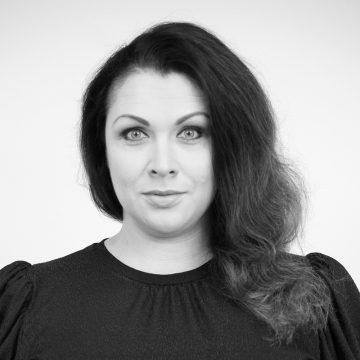 Mustavalkoinen muotokuva naisesta, jolla on pitkät, aaltoilevat tummat hiukset, yllään tumma toppi, katselee suoraan kameraan neutraali ilme tavallista taustaa vasten.