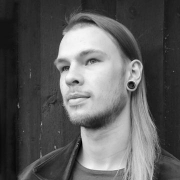 Mustavalkoinen muotokuva nuoresta henkilöstä, jolla on pitkät vaaleat hiukset, parta ja suuret pistokekorvakorut ja joka katsoo mietteliäästi sivulle. Taustalla on tumma puinen seinä.