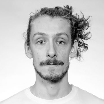 Mustavalkoinen muotokuva nuoresta miehestä, jolla on viikset ja parta, yllään vaalea paita. Hänen aaltoilevat hiuksensa on sidottu yhteen sivuun. Hän katsoo suoraan kameraan neutraali ilme kasvoillaan.