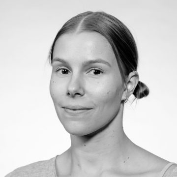 Mustavalkoinen muotokuva nuoresta naisesta, jolla on vaalea iho ja suorat hiukset, jotka on erotettu keskeltä ja vedetty taakse matalaksi ponnariksi, päällään vaalea toppi ja pienet korvarenkaat, katsoo kameraan.