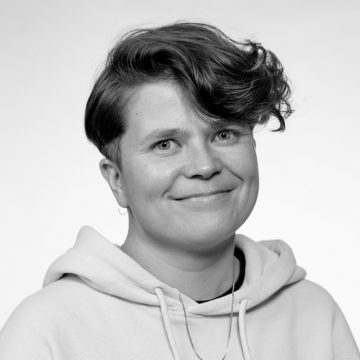 Mustavalkoinen muotokuva henkilöstä, jolla on lyhyet, aaltoilevat hiukset, päällään vaalea huppari ja hento hymy, katselee hieman sivulle tavallista taustaa vasten.