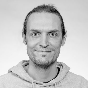 Mies, jolla on pitkät taakse vedetyt hiukset ja vaalea huppari, hymyilee hieman kameralle. Tausta on tavallinen ja vaalea, ja kuva on mustavalkoinen.
