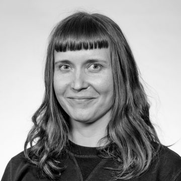 Nainen, jolla on suorat, olkapään pituiset hiukset ja tylppä otsatukka ja jolla on tumma toppi, hymyilee kameralle pehmeästi vaaleaa taustaa vasten. Kuva on mustavalkoinen.