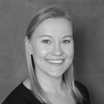 Mustavalkoinen muotokuva hymyilevästä naisesta, jolla on suorat, vaaleat hiukset ja tumma toppi, yksivärisen taustan edessä.