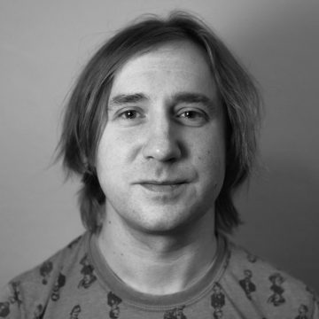 Henkilö, jolla on suorat, olkapäiden pituiset hiukset, katsoo kameraan hymyillen. Hänellä on yllään kuviollinen paita. Kuva on mustavalkoinen ja siinä on neutraali tausta.