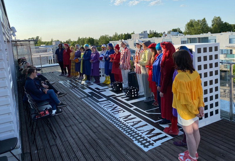 Ryhmä värikkäisiin pukuihin pukeutuneita esiintyjiä seisoo rivissä ulkokattolavalla, ja yleisö istuu vasemmalla. Lavalla on kaupunkikatu, johon on kirjoitettu ZONE MALMI. Taustalla näkyy moderneja rakennuksia.