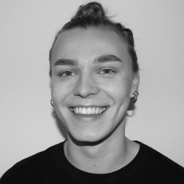 Henkilö, jolla on pitkät taakse sidotut hiukset, korvakorut ja musta toppi, hymyilee kameralle vaaleaa taustaa vasten. Kuva on mustavalkoinen.