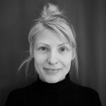 Nainen, jolla on vaaleat vaaleat hiukset sotkuisessa pussissa, hymyilee pehmeästi kameralle. Hänellä on yllään musta poolopusero, ja hän on kuvassa tummaa taustaa vasten. Kuva on mustavalkoinen.
