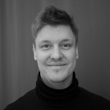 Nuori mies, jolla on lyhyet, vaaleat hiukset ja vaaleat kasvokarvat, hymyilee pehmeästi. Hänellä on yllään musta poolopusero, ja hänet on kuvattu mustavalkoisena tavallista, sumeaa taustaa vasten.