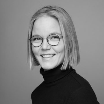 Nuorella naisella, jolla on suorat, leuan pituiset vaaleat hiukset, on pyöreät silmälasit ja musta poolopusero. Hän hymyilee ja katsoo hieman sivulle vaaleanharmaata taustaa vasten. Kuva on mustavalkoinen.