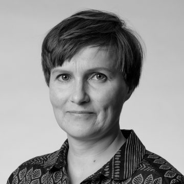 Mustavalkoinen muotokuva henkilöstä, jolla on lyhyet hiukset, päällään kuviollinen paita, katselee kameraan neutraali ilme tavallista taustaa vasten.