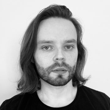 Henkilö, jolla on olkapäiden pituinen tukka ja parta, katsoo kameraan neutraalilla ilmeellä, ja hänellä on musta paita vaaleaa taustaa vasten.