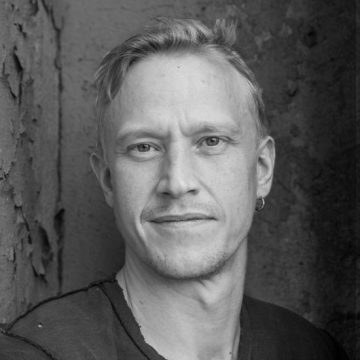 Mustavalkoinen muotokuva lyhyet vaaleat hiukset omaavasta miehestä, jolla on tumma paita ja pieni korvarengas, seisoo kuvioitua, kuorittua seinää vasten ja katsoo suoraan kameraan neutraalilla ilmeellä.