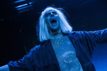 Vaaleansininen esiintyjä, jolla on vaaleansininen iho, valkoinen peruukki ja tumma hapsupuku, seisoo dramaattisen sinisen näyttämövalaistuksen alla, suu auki kuin laulaisi tai huutaisi, kädet ojennettuina.