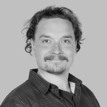 Vaaleaihoinen mies, jolla on aaltoilevat ruskeat hiukset, viikset ja parta, hymyilee hieman. Hänellä on ruudullinen paita t-paidan päällä, ja hän poseeraa vaaleanharmaata taustaa vasten.
