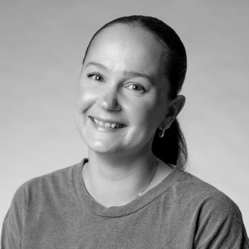 Nainen, jolla on tummat hiukset taaksepäin vedettynä, hymyilee ja jolla on yllään tavallinen t-paita. Kuva on mustavalkoinen, ja taustalla on neutraali tausta.