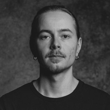 Vaaleaihoinen, viiksekäs ja pukinpartainen nuori henkilö tuijottaa suoraan kameraan. Hänellä on pitkät hiukset taaksepäin vedettynä, hänellä on rengaskorvakorut ja tumma paita. Taustalla on kuvioitu tumma pinta. Kuva on mustavalkoinen.