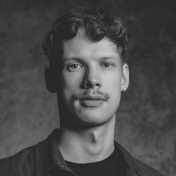 Mustavalkoinen muotokuva nuoresta miehestä, jolla on lyhyet kiharat hiukset, viikset ja pieni korvakoru vasemmassa korvassa. Hänellä on kauluspaita t-paidan päällä ja hän katsoo suoraan kameraan.