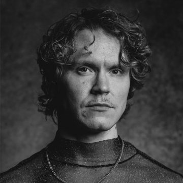 Mustavalkoinen muotokuva henkilöstä, jolla on aaltoileva tukka, hennot viikset ja vakava ilme, yllään hohtava korkeakaulainen paita ja ketjukoru tummaa, kuvioitua taustaa vasten.