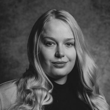 Mustavalkoinen muotokuva naisesta, jolla on pitkät, vaaleat, aaltoilevat hiukset, päällään tumma poolokaulus ja vaalea takki, katselee kameraan hymyillen kuvioitua taustaa vasten.