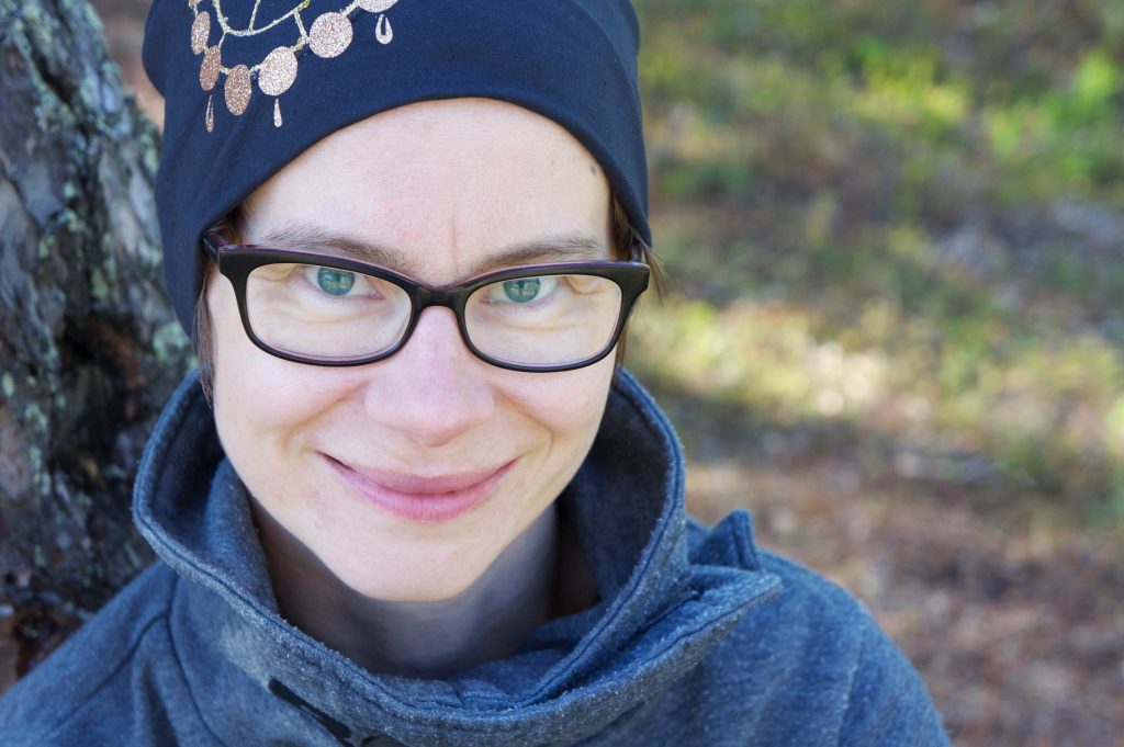 Henkilö, jolla on silmälasit, tumma koristeellinen huivi ja vetoketjullinen takki, hymyilee nojaillessaan puuhun ulkona. Tausta on sumea, ja siinä näkyy vehreyttä ja auringonvaloa.