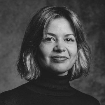 Mustavalkoinen muotokuva naisesta, jolla on leuan pituiset hiukset, päällään tumma poolopusero, katselee kameraan hento hymyillen. Tausta on pehmeästi sumea.