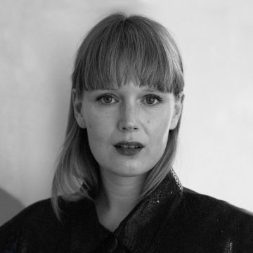 Mustavalkoinen muotokuva naisesta, jolla on suorat, vaaleat hiukset ja otsatukka ja joka katsoo suoraan kameraan neutraalilla ilmeellä. Hänellä on yllään kiiltävä tumma toppi. Tausta on tavallinen ja pehmeästi valaistu.