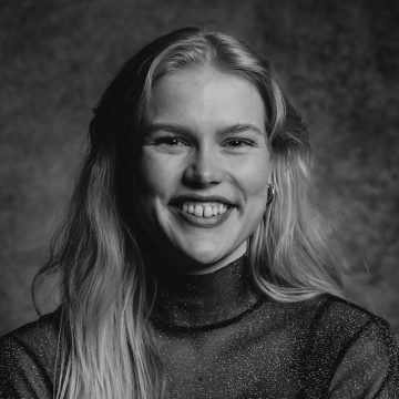 Mustavalkoinen muotokuva nuoresta naisesta, jolla on pitkät vaaleat hiukset ja joka hymyilee kameralle. Hänellä on yllään läpikuultava, kimalteleva toppi, ja tausta on pehmeästi sumea.
