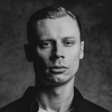 Mustavalkoinen muotokuva nuoresta miehestä, jolla on lyhyet, siististi muotoillut hiukset ja joka katsoo suoraan kameraan neutraali ilme kasvoillaan. Hänellä on yllään tumma paita vaalean t-paidan päällä, ja tausta on pehmeästi sumea.