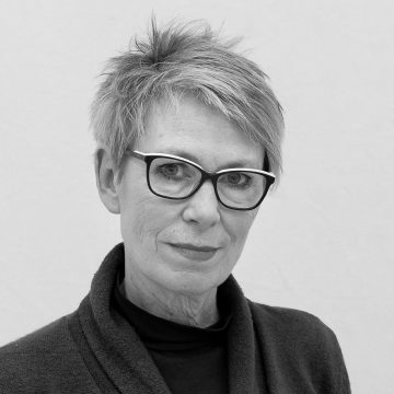 Lyhyet, vaaleat hiukset ja silmälasit omaavalla naisella on yllään tumma villapaita, ja hän katsoo kameraan neutraalilla ilmeellä tavallista taustaa vasten.