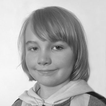 Mustavalkoisessa muotokuvassa on lapsi, jolla on suorat, vaaleat hiukset ja hymyilevä huppari, ja joka on kuvattu tavallista vaaleaa taustaa vasten.