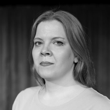 Nainen, jolla on pitkät, suorat hiukset, katsoo rauhallisesti kameraan. Hänellä on yllään vaalea toppi. Tausta on sumea, ja kuva on mustavalkoinen.