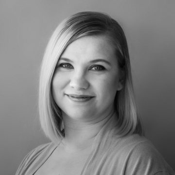 Mustavalkoinen muotokuva naisesta, jolla on suorat, hartioiden pituiset vaaleat hiukset, yllään vaalea toppi ja joka hymyilee pehmeästi katsoen kameraan tavallista taustaa vasten.