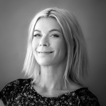 Mustavalkoinen muotokuva naisesta, jolla on vaaleat, olkapään pituiset hiukset, yllään kukkatoppi, hymyilee kevyesti ja katsoo kameraan tavallista taustaa vasten.