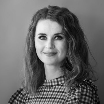 Mustavalkoinen muotokuva naisesta, jolla on aaltoileva tukka, päällään ruudullinen toppi, jossa on röyhelöiset olkapäät, hymyilee hieman ja katsoo kameraan tavallista taustaa vasten.