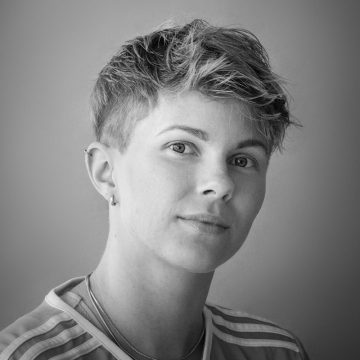 Mustavalkoinen muotokuva henkilöstä, jolla on lyhyet, kuvioidut hiukset ja alaleikkaus, päällään raidallinen paita ja pienet korvarenkaat, katselee suoraan kameraan neutraali ilme.