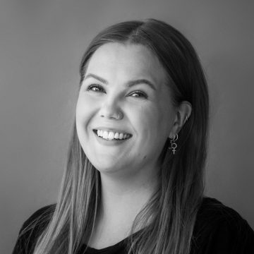 Nuori pitkätukkainen nainen hymyilee kameralle. Hänellä on nenäsormus, kaksi korvakorua, joissa on ankh-symboli, ja tumma toppi. Tausta on yksinkertainen ja neutraali. Kuva on mustavalkoinen.