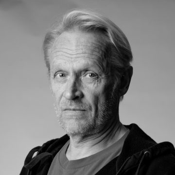 Mustavalkoinen muotokuva vanhemmasta miehestä, jolla on vaaleat hiukset ja lyhyt parta, yllään rento paita ja takki, ja joka katsoo vakavasti kameraan tavallista taustaa vasten.
