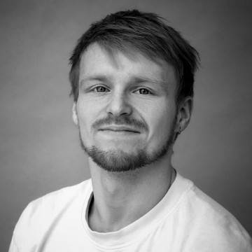 Mustavalkoinen muotokuva nuoresta miehestä, jolla on lyhyet hiukset, leikattu parta ja pieni korvakoru. Hänellä on yllään tavallinen vaalea t-paita ja hän hymyilee hieman katsoen kameraan. Tausta on tavallinen.