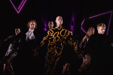 Ryhmä dramaattisiin pukuihin pukeutuneita esiintyjiä poseeraa pimeällä näyttämöllä, jonka taustalla on violetti valaistus ja neonvärisiä muotoja, ja katsoo intensiivisesti kohti yleisöä.