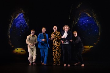 Viisi teatteripukuista miestä seisoo näyttämöllä dramaattisissa asennoissa, ja taustalla on kaksi luolan sisäänkäyntiä, joista näkyy yötaivas, jolla on puita ja tähtiä.