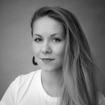Mustavalkoinen muotokuva nuoresta naisesta, jolla on pitkät vaaleat hiukset, valkoinen paita, korvakorut ja nenärengas, ja joka katsoo kameraan neutraali ilme kasvoillaan tavallista taustaa vasten.